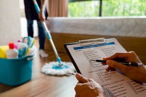 Person holding a cleaning checklist while another person mops the floor in the background.