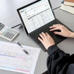 Person typing on a laptop with a spreadsheet and calculator on the desk.