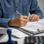 Man signing a document at a desk with a laptop and calculator