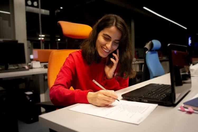 A woman sits at a desk with a laptop and phone, managing time zones for her work tasks.