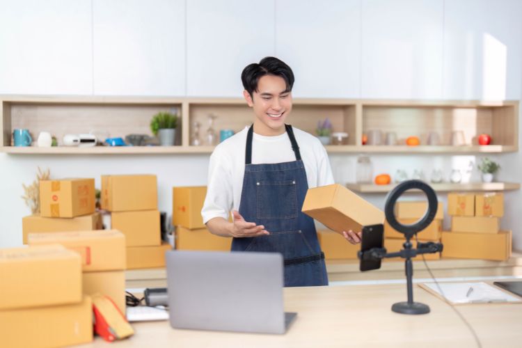 A man wearing an apron stands with a laptop and boxes, indicating he is engaged in a work-related activity.