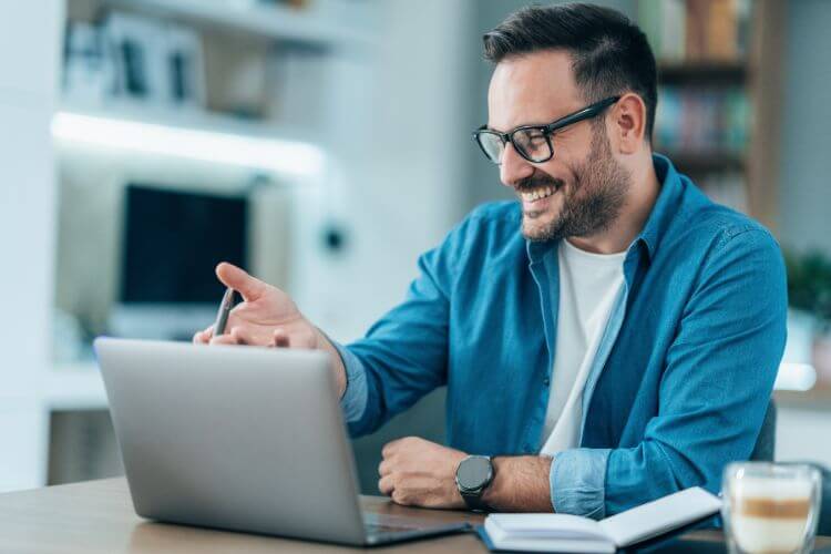 A man with glasses smiles while working on his laptop, appearing focused and content.