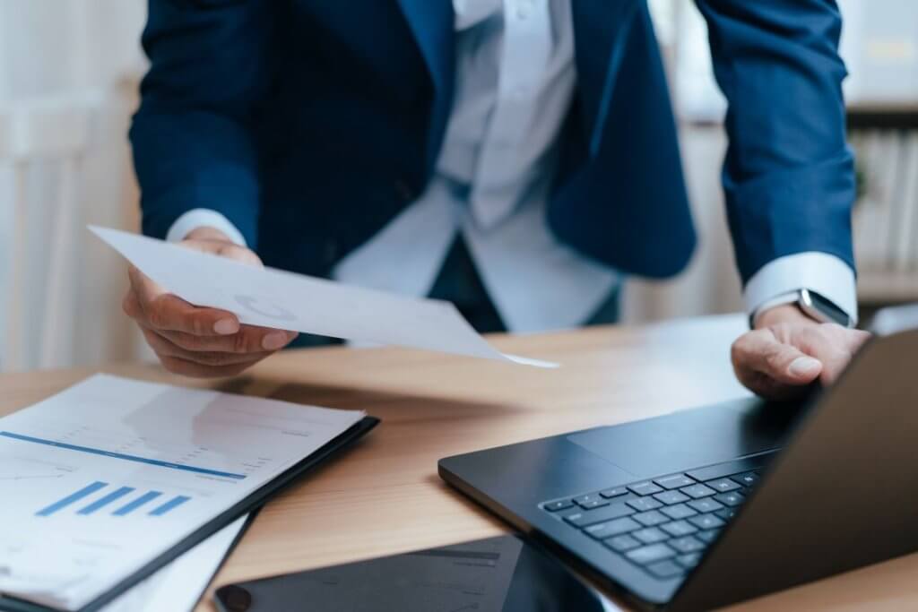 Business professional reviewing reports and documents while working on a laptop.