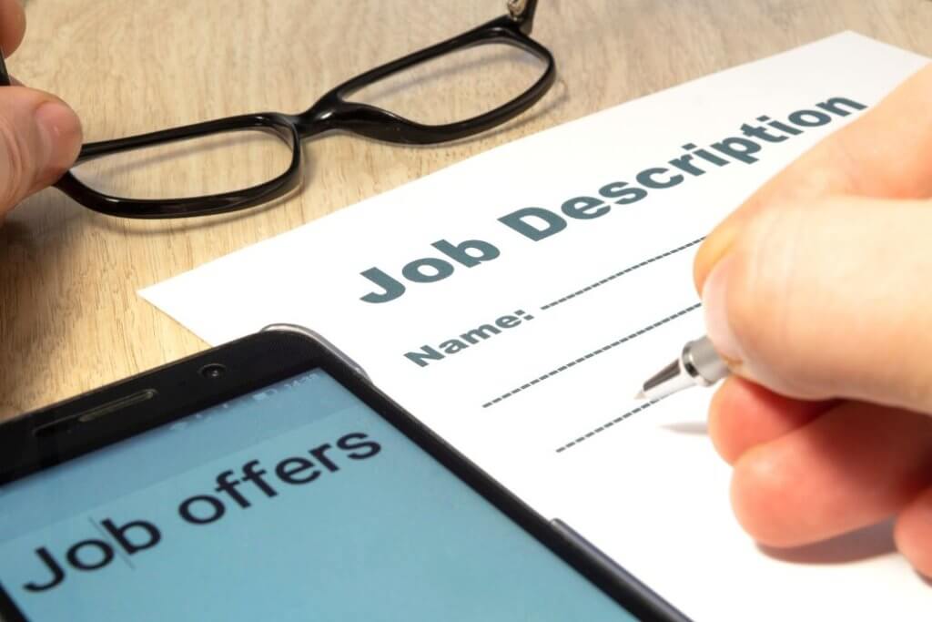 Person completing a job description beside a phone showing job offers.