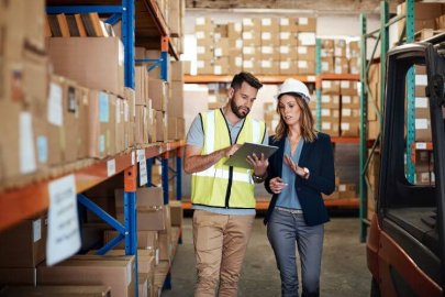 A man in a safety vest discussing with a woman in a hard hat inside a warehouse filled with boxes