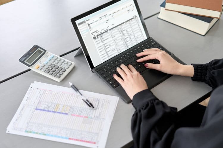 Person typing on a laptop with a spreadsheet and calculator on the desk.